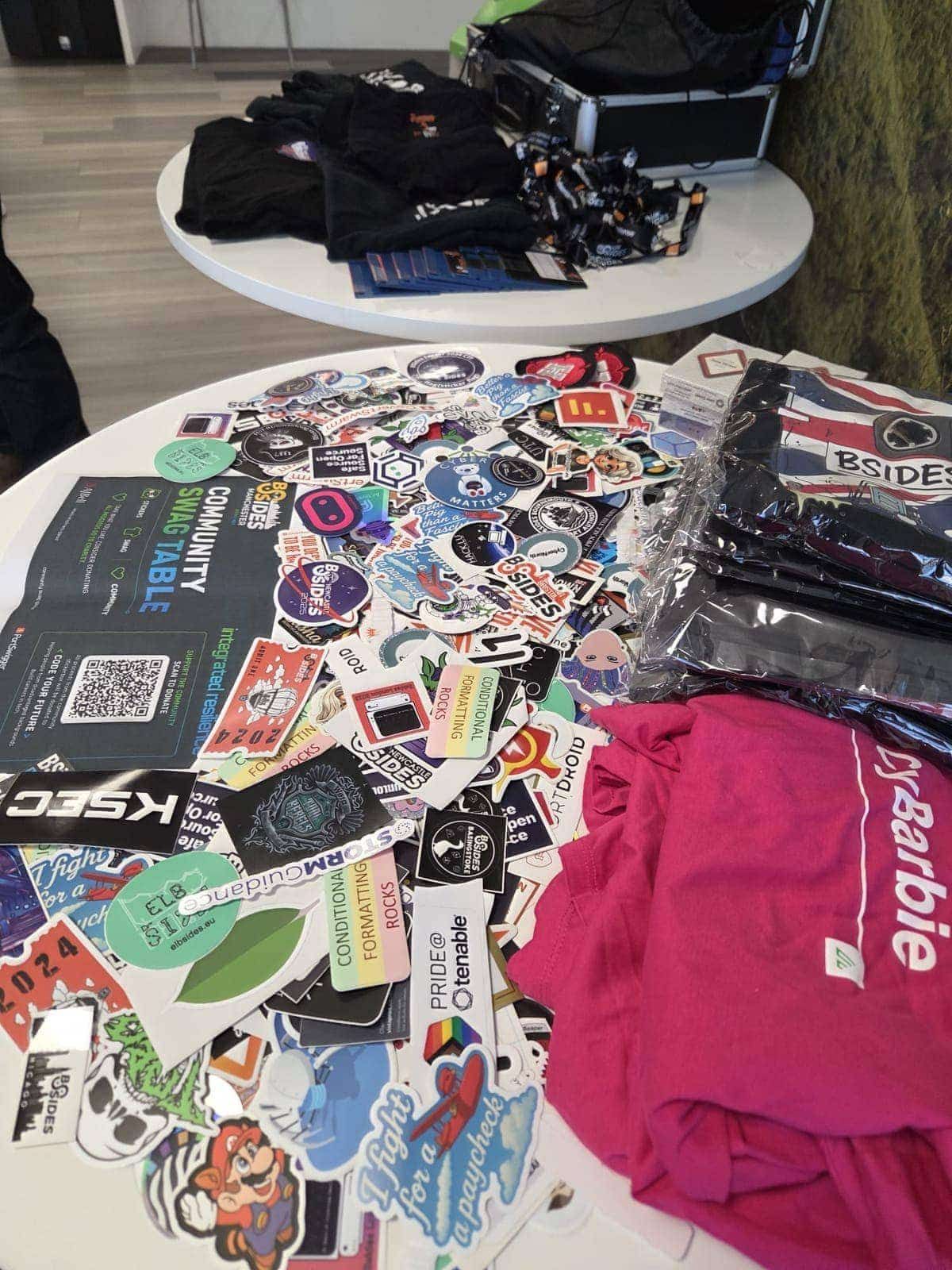 A close-up of the community swag table, with stickers from a wide range of community projects and BSides Manchester t-shirts