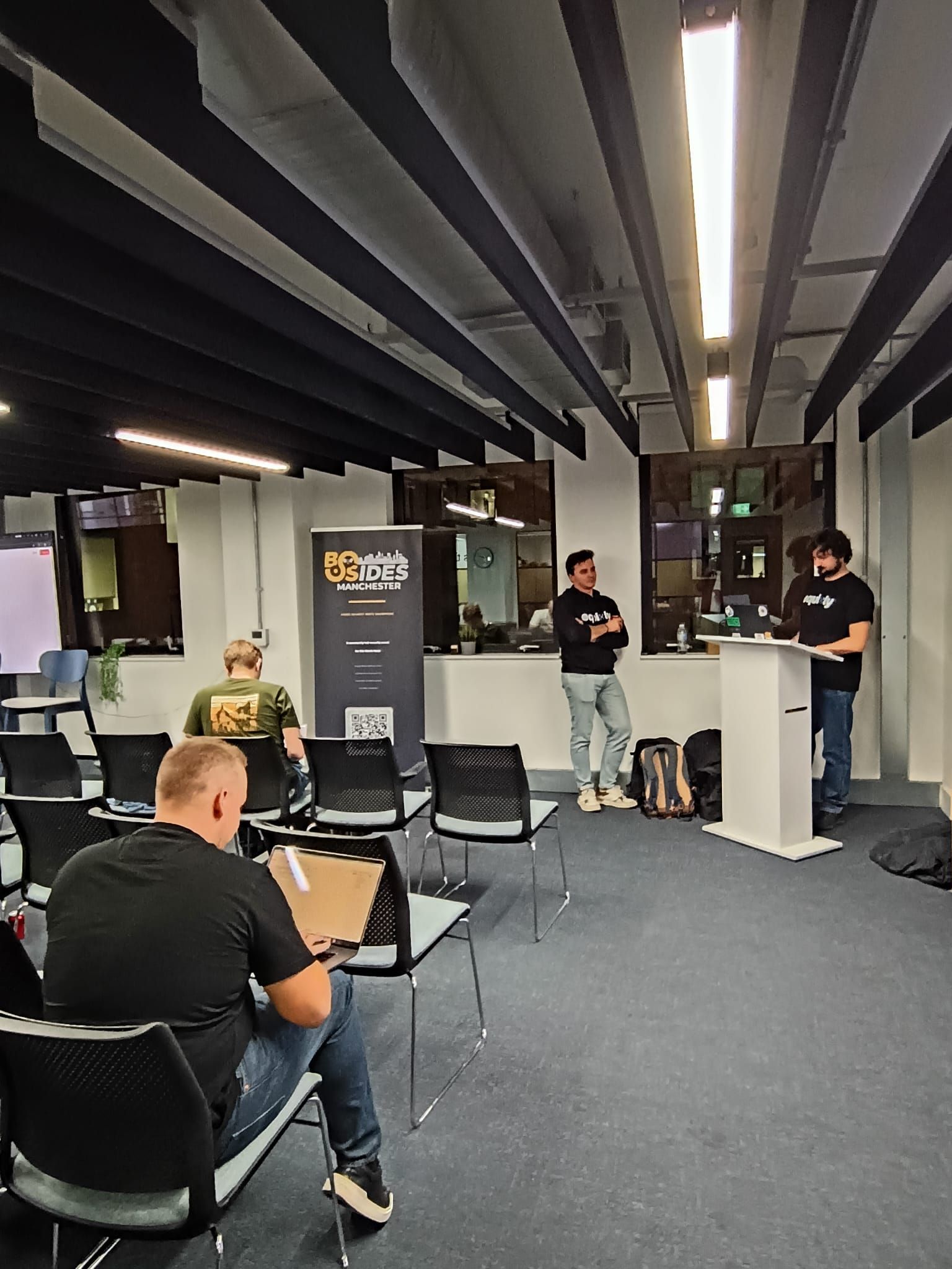 Two members of the Equixly team at the podium introducing the live CTF, next to the BSides Manchester roller banner