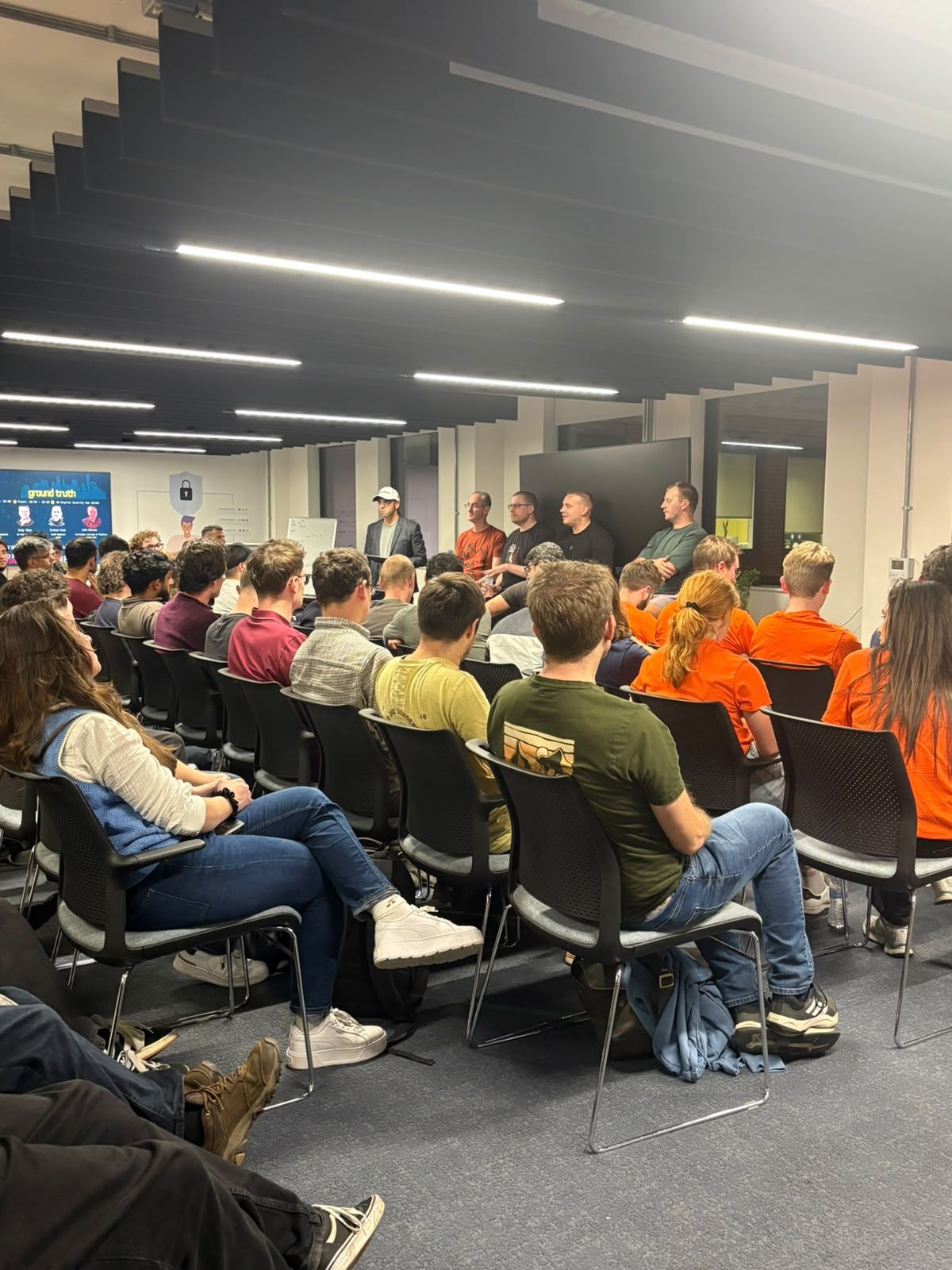 The audience at DiSH, mostly seen from behind, facing the moderator and four Ground Truth panellists, with the Ground Truth title slide on a screen to the left