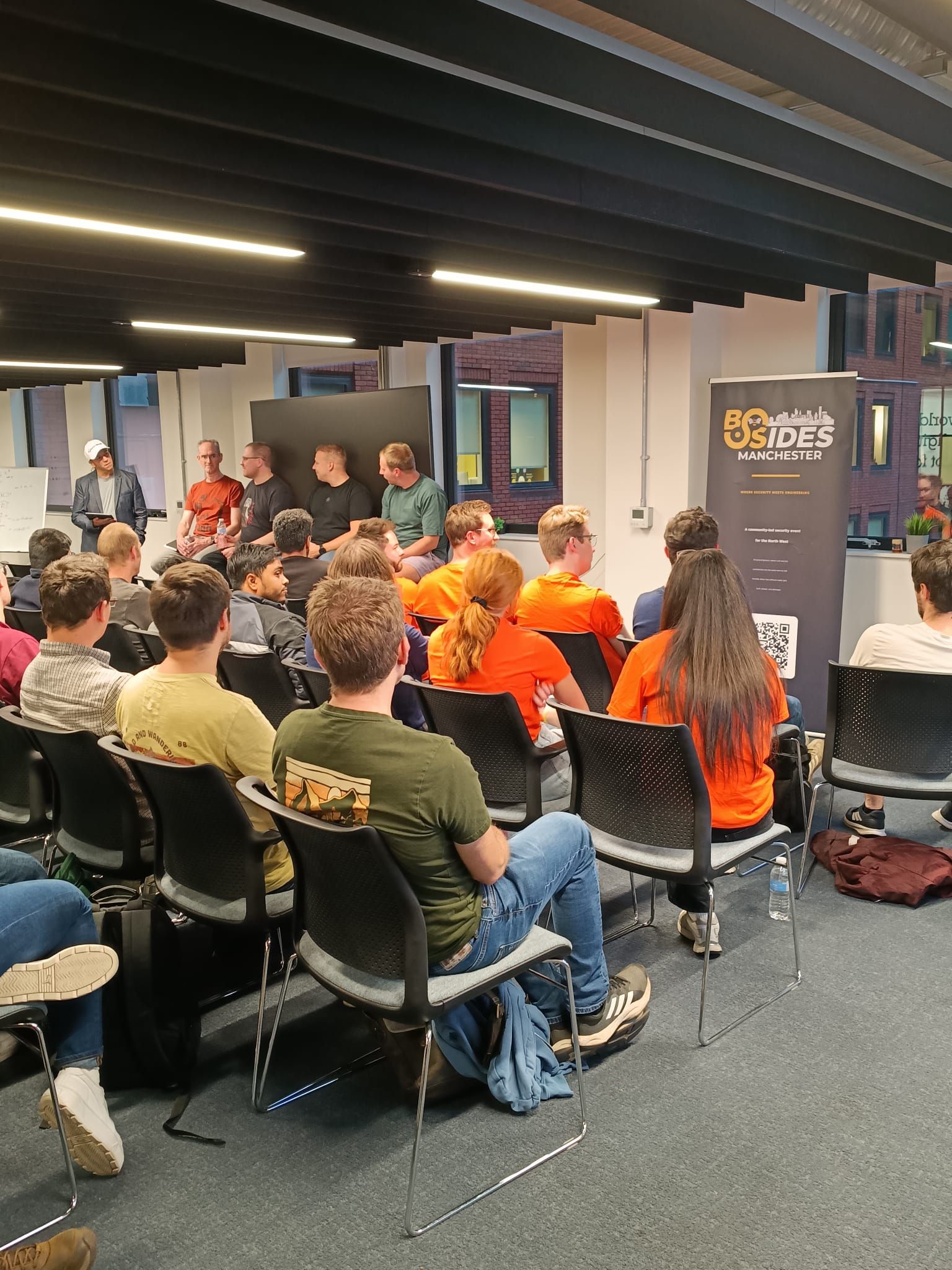 The room at DiSH during the Ground Truth panel, seen from behind the seated audience, with the moderator and four panellists at the front and the BSides Manchester roller banner on the right